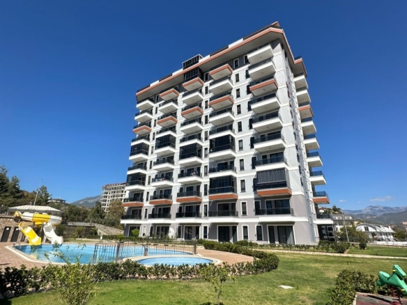 Exterior view of Bravo Tower residential complex in Demirtas, Alanya. Showing a modern multi-story apartment building, outdoor swimming pool with water slide, green landscaped garden, and children's playground area.