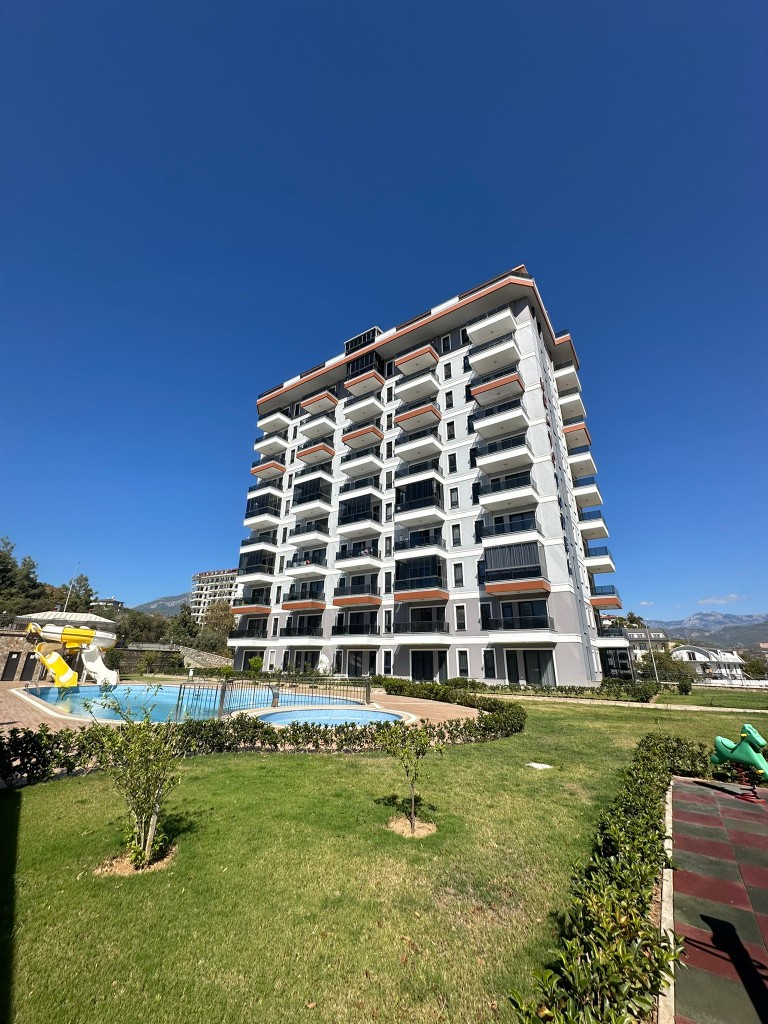 Exterior view of Bravo Tower residential complex in Demirtas, Alanya. Showing a modern multi-story apartment building, outdoor swimming pool with water slide, green landscaped garden, and children's playground area.