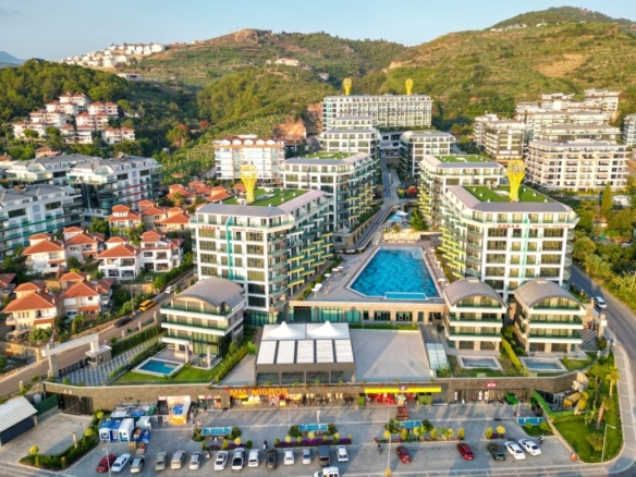 Вид с дрона на прибрежный жилой комплекс «Konak Seaside Premium», выставленный на продажу в Каргыджаке, Аланья. Комплекс состоит из 8 корпусов, имеет большой центральный бассейн, ландшафтные сады, а также торговые помещения на первом этаже, в том числе магазины «Migros» и «BİM». На заднем плане видны горы Тавр.
