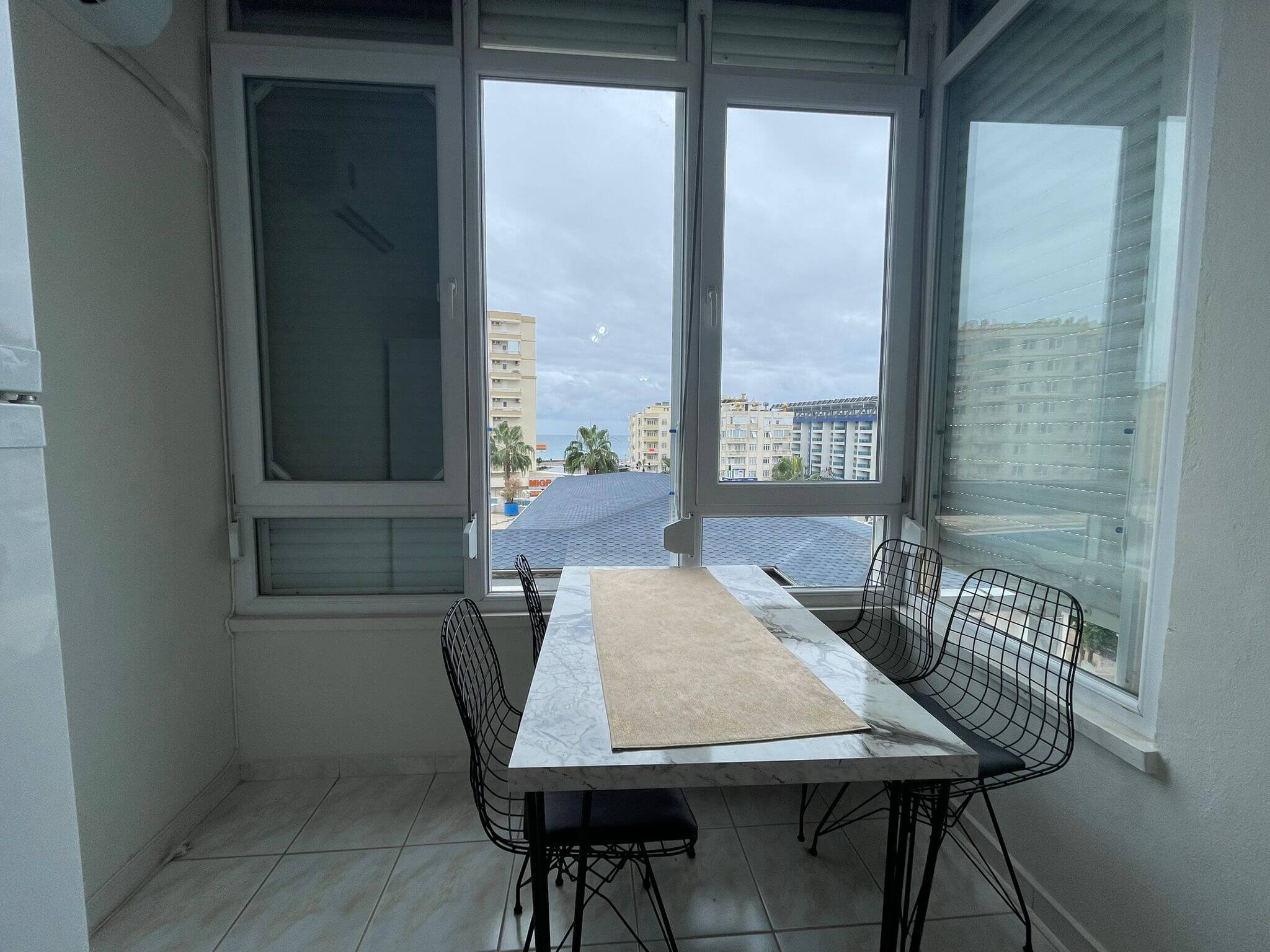 A bright enclosed dining area featuring a marble-look rectangular table with four black wire-frame chairs. Large floor-to-ceiling windows provide natural light and overlook nearby apartment buildings, palm trees, and a distant sea view beneath a cloudy sky. The space has white walls, tiled flooring, and a clean, modern design.
