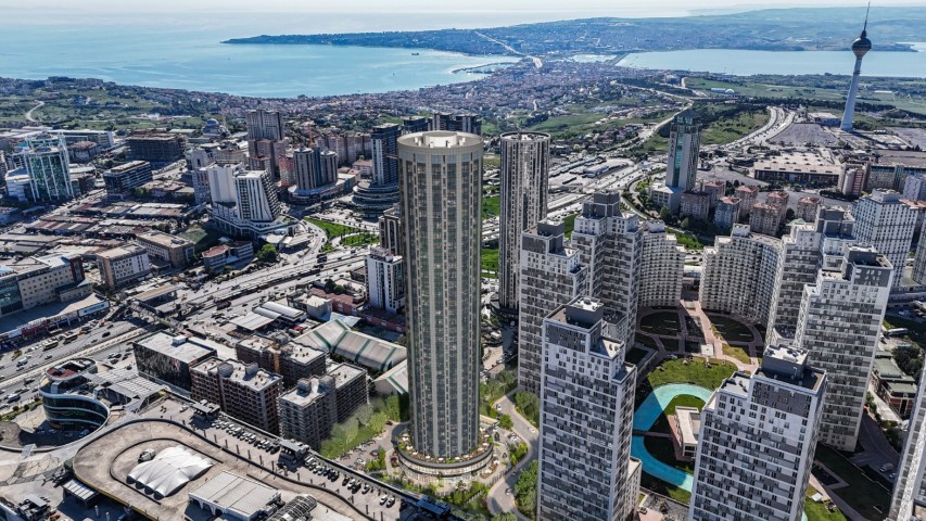 An aerial drone photograph looking towards the Marmara Sea, showcasing a vast urban landscape. The central focus is the towering, modern, cylindrical Kristal Kule skyscraper, defined by its glass and metal facade and distinct crown. Adjacent to it are interconnected, multi-story, stepped residential blocks. The complex features manicured green spaces and a water feature. An intricate road network with cars surrounds the development, and in the far background, a prominent communication and TV tower stands against the blue sea and distant hills. A cinematic black bar with white text is present at the bottom of the frame.