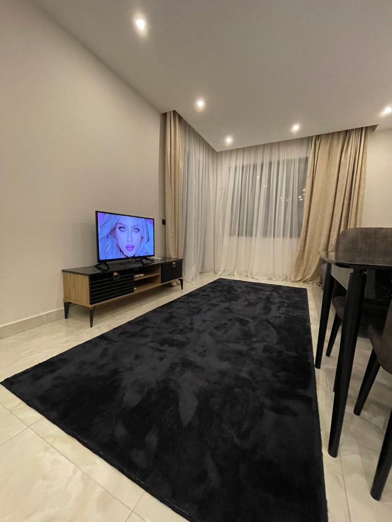 Living room interior showing a flat-screen TV on a wooden stand, a large black rug, and a portion of the dining area