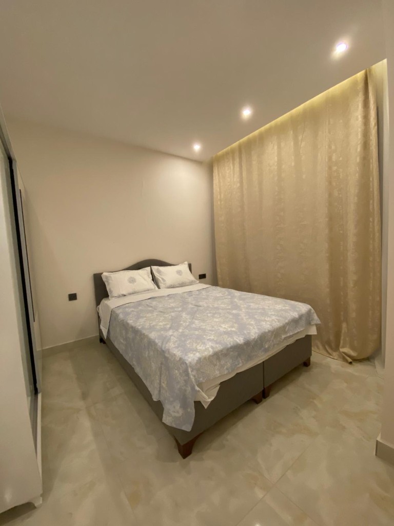 Bedroom interior showing a grey upholstered double bed and long beige curtains under recessed ceiling lights