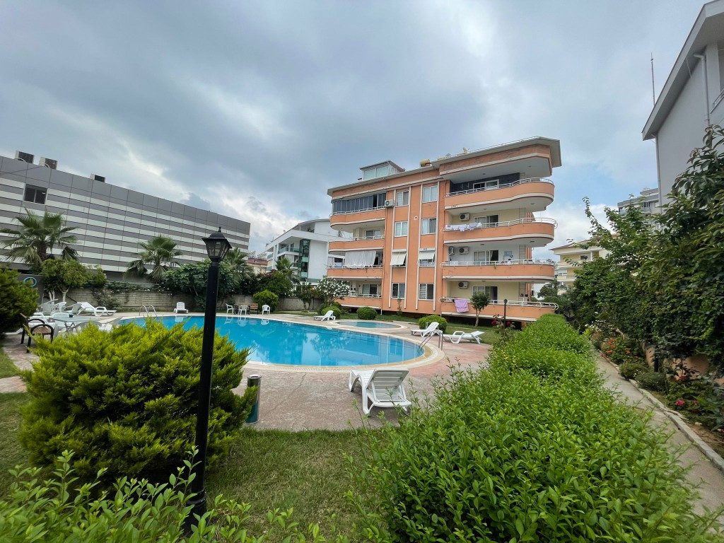 Exterior view of Tara Sitesi residential building in Oba Alanya featuring a curved outdoor swimming pool and sun loungers surrounded by green garden landscaping
