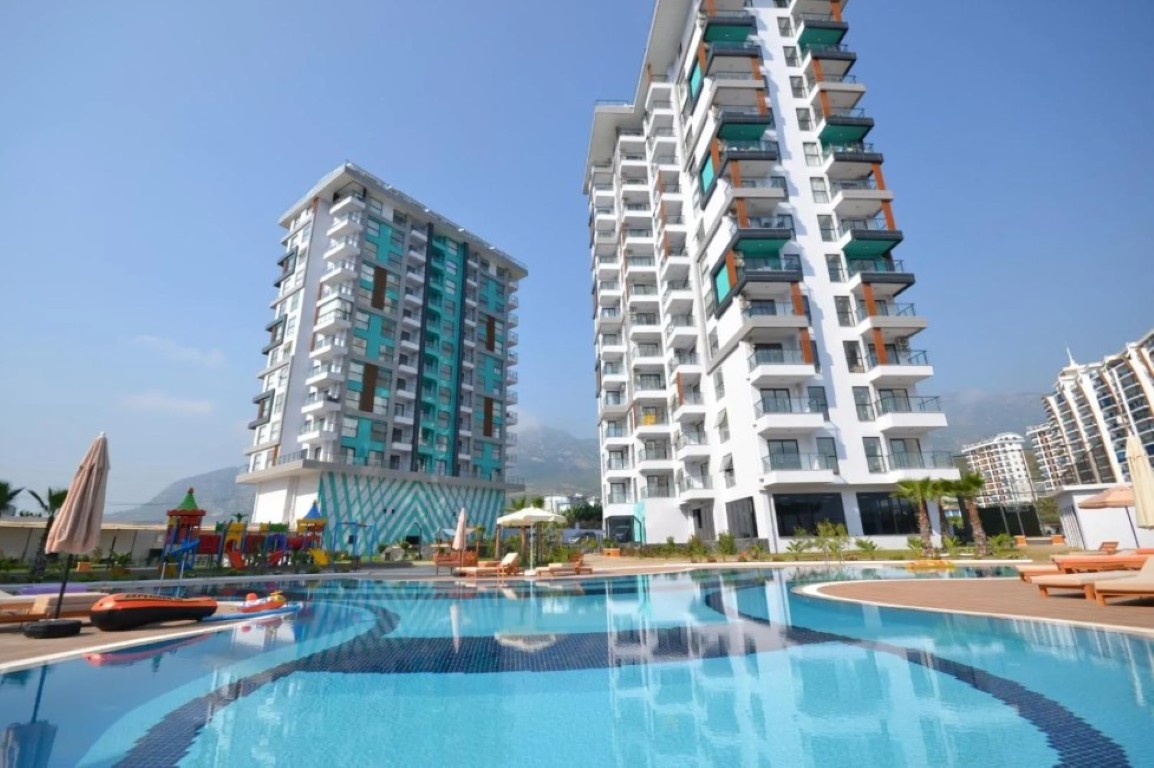Exterior view of State of Art Residence in Mahmutlar featuring a large outdoor swimming pool, sun loungers, children's playground, and modern high-rise blocks.