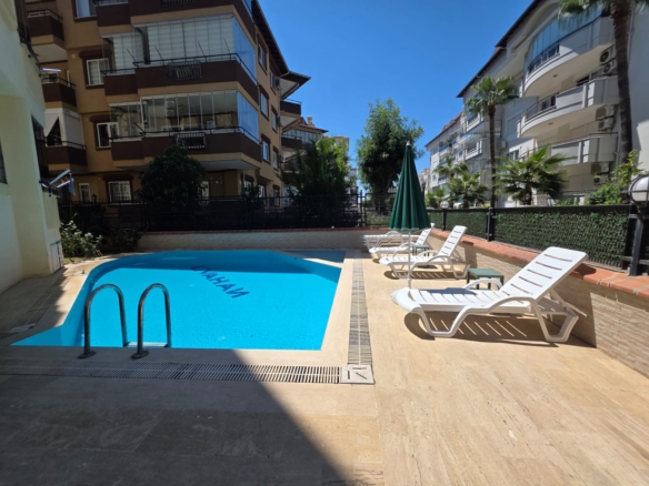 Utomhuspool vid bostadskomplexet Livahan, med klart blått vatten och ordet ”LIVAHAN” på poolbotten, metallräcken, vita solstolar i plast, ett stängt grönt parasoll, kaklat däck, palmer och flervåningshus under en klarblå himmel.
