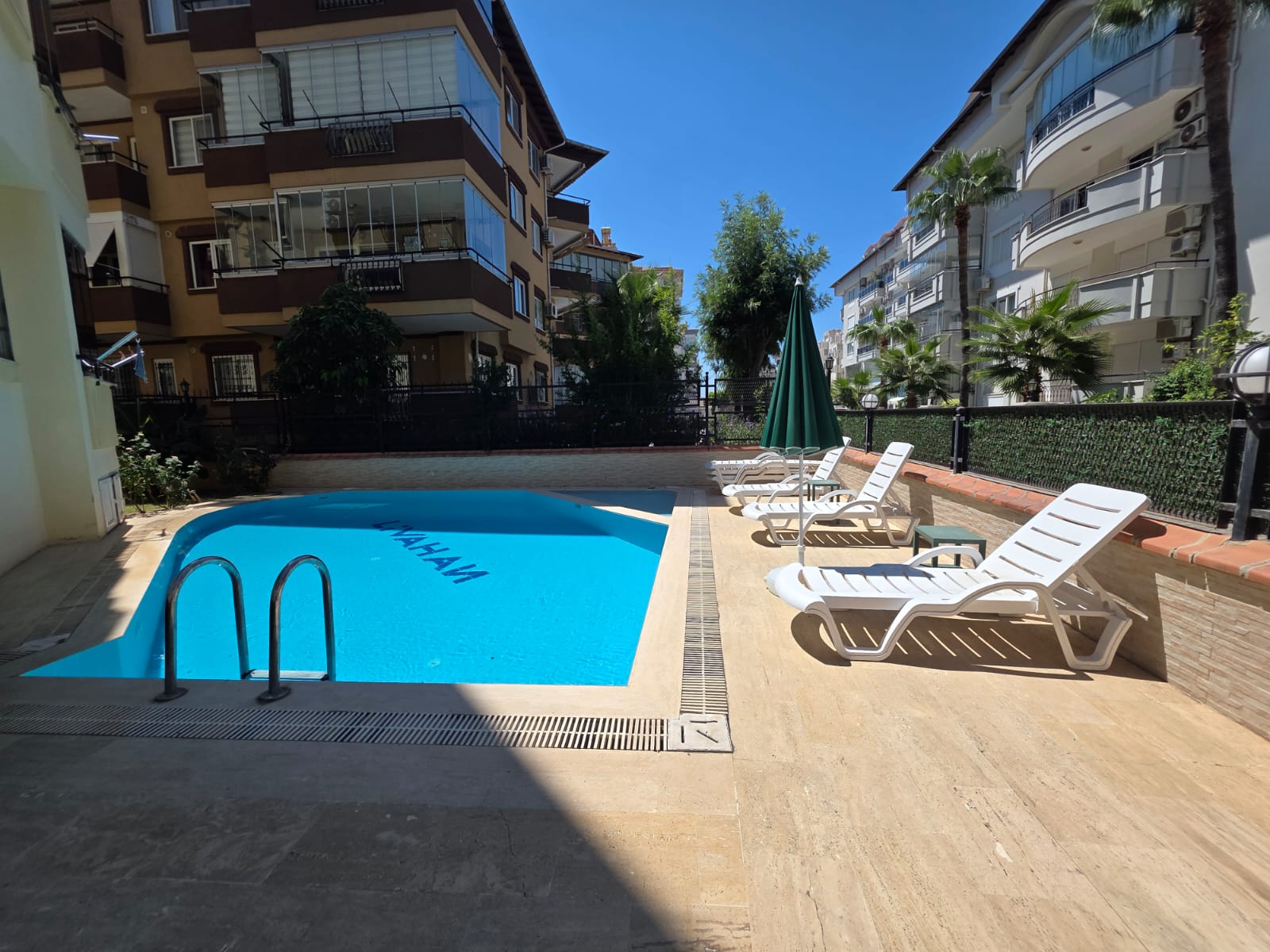Outdoor swimming pool at the Livahan residential complex, featuring clear blue water with the word “LIVAHAN” on the pool floor, metal handrails, white plastic sun loungers, a closed green parasol, tiled deck, palm trees, and multi-story apartment buildings under a bright blue sky.